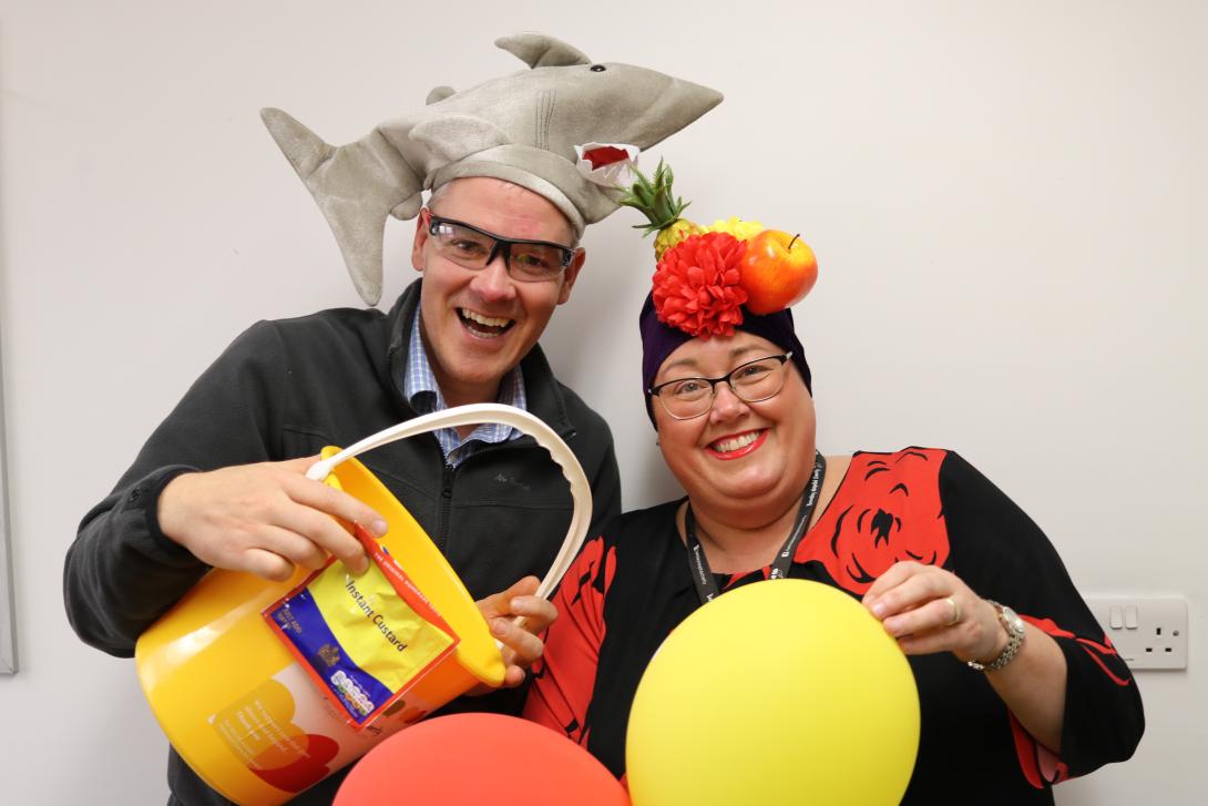 Two volunteers for Barnsley Hospital charity, smiling - one person is wearing a hat in the shape of a shark, and holds a bucket. The other person is holding orange and yellow balloons. 