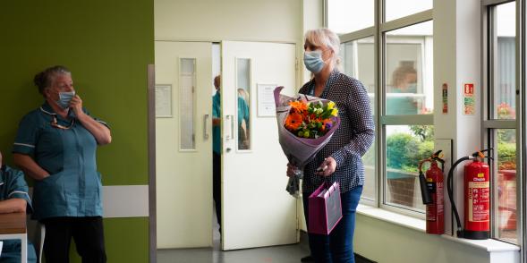 A woman with white hair tied up, wearing a lilac jumper and blue jeans arrives to the hospital with a bunch of flowers.