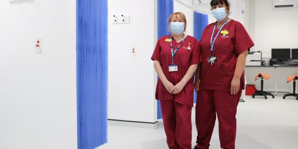 Two healthcare assistants in red scrubs, smile from behind masks, whilst standing in a clinical area of the CDC