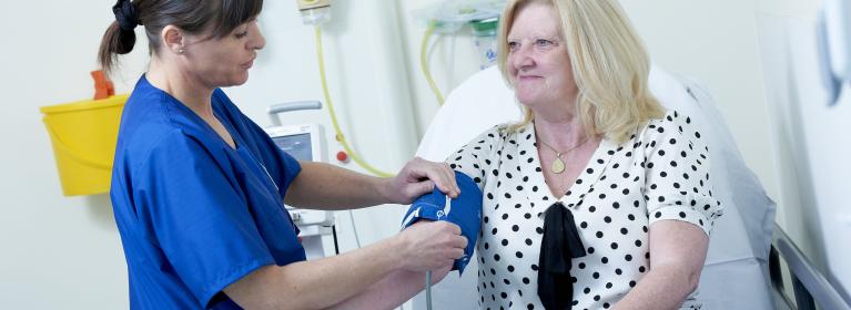 A nurse in blue scrubs who is white with brown hair, takes the blood pressure of a patient. The patient is a white woman with blonde hair. She is smiling at the nurse.