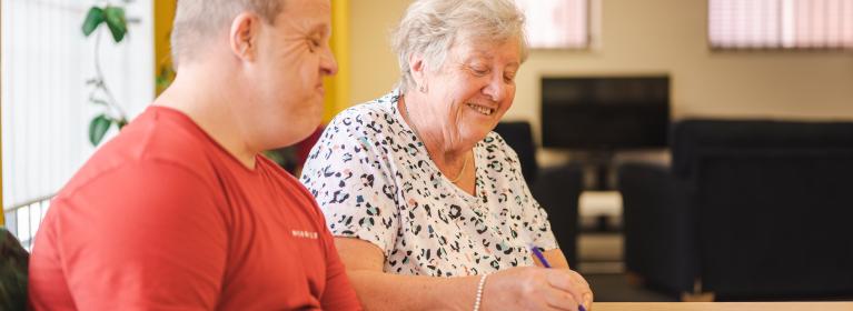 An person of "mid-age" looks on, as an older person smiles, whilst colouring in