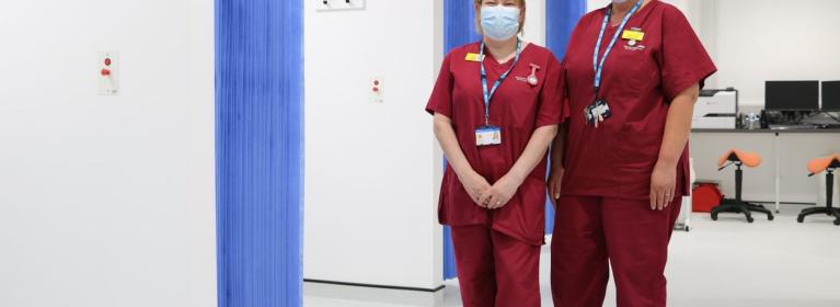 Two healthcare assistants in red scrubs, smile from behind masks, whilst standing in a clinical area of the CDC