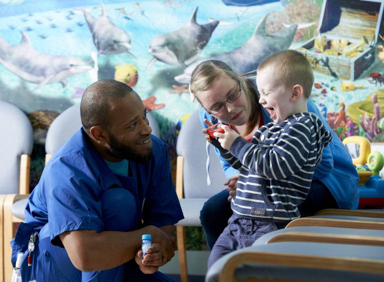 A nurse who is a Black man smiles at a young boy who is his patient; the boy is laughing. His mum sits beside him. 