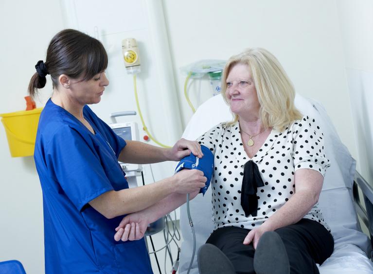 A nurse in blue scrubs who is white with brown hair, takes the blood pressure of a patient. The patient is a white woman with blonde hair. She is smiling at the nurse.