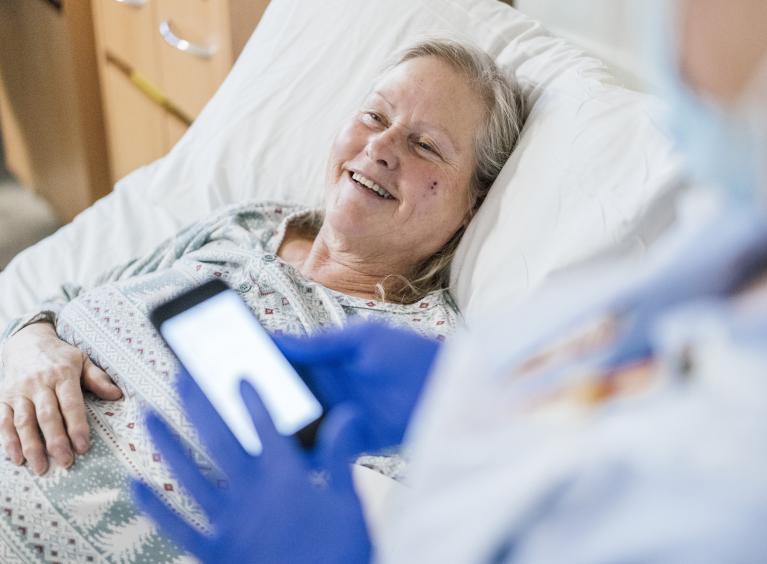An older patient smiles as a member of staff uses a digital device to record healthcare information