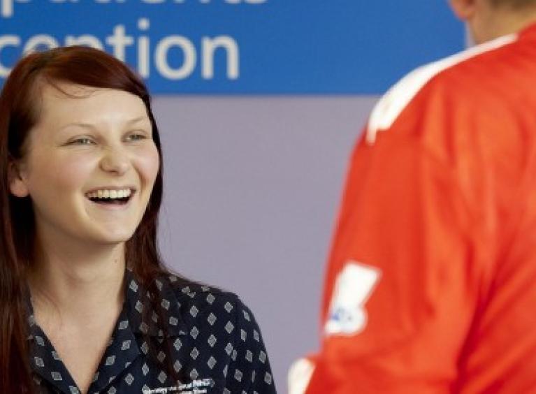 A receptionist smiles and laughs as she welcomes a patient to Barnsley Hospital. She is wearing a navy blouse and has long copper hair.