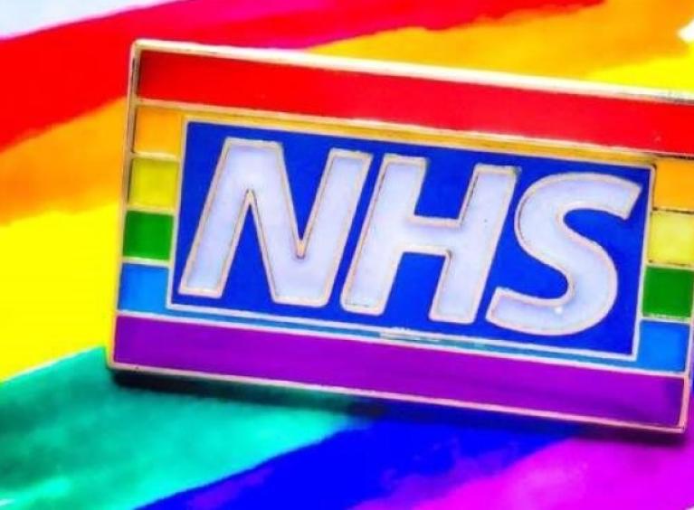 A photo of the pro LGBT+ inclusion "NHS Rainbow badge" - which is an enamel badge showing the NHS logo in front of a horizontal rainbow coloured flag - sitting atop a rainbow coloured painting.