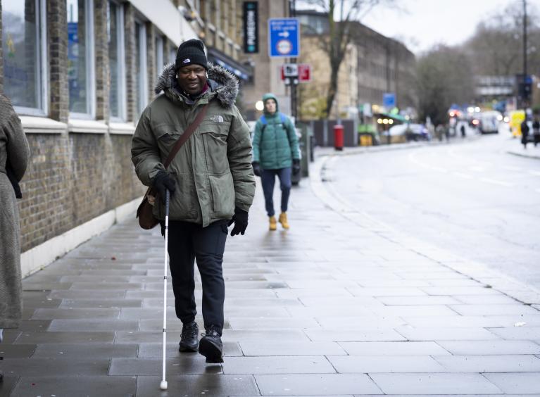 An older man walks up a high street whilst using a cane.