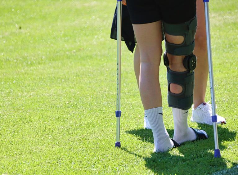 A young person with a leg injury walks across a park, assisted by another person, wearing a leg brace and using crutches.