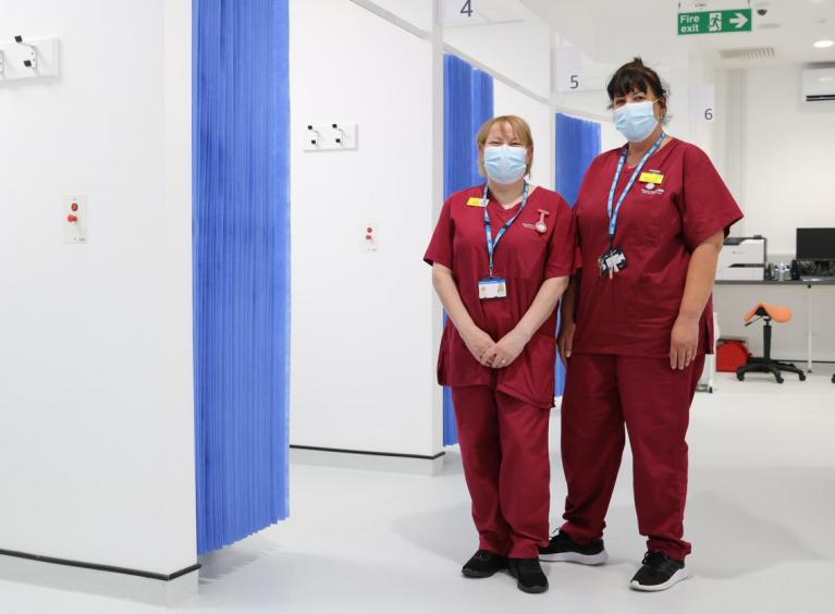 Two healthcare assistants in red scrubs, smile from behind masks, whilst standing in a clinical area of the CDC