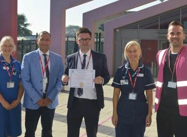 Four people stand outside a school as a doctor presents a certificate to the school's principal; they are joined by two nurses and a teacher 