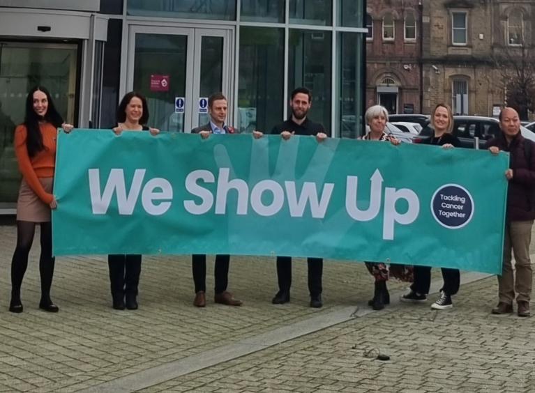 Seven people holding a green banner. The banner says we show up in white text.