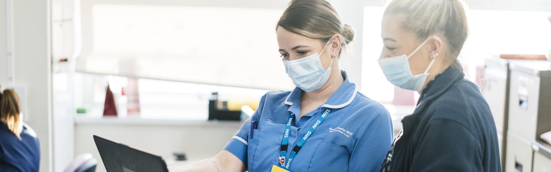 Two nurses stand looking at the screen of a laptop