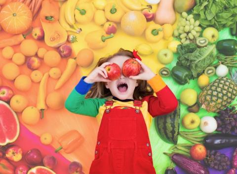 A young girl holds two apples in front of her eyes, standing in front of a rainbow of fruit and vegetables