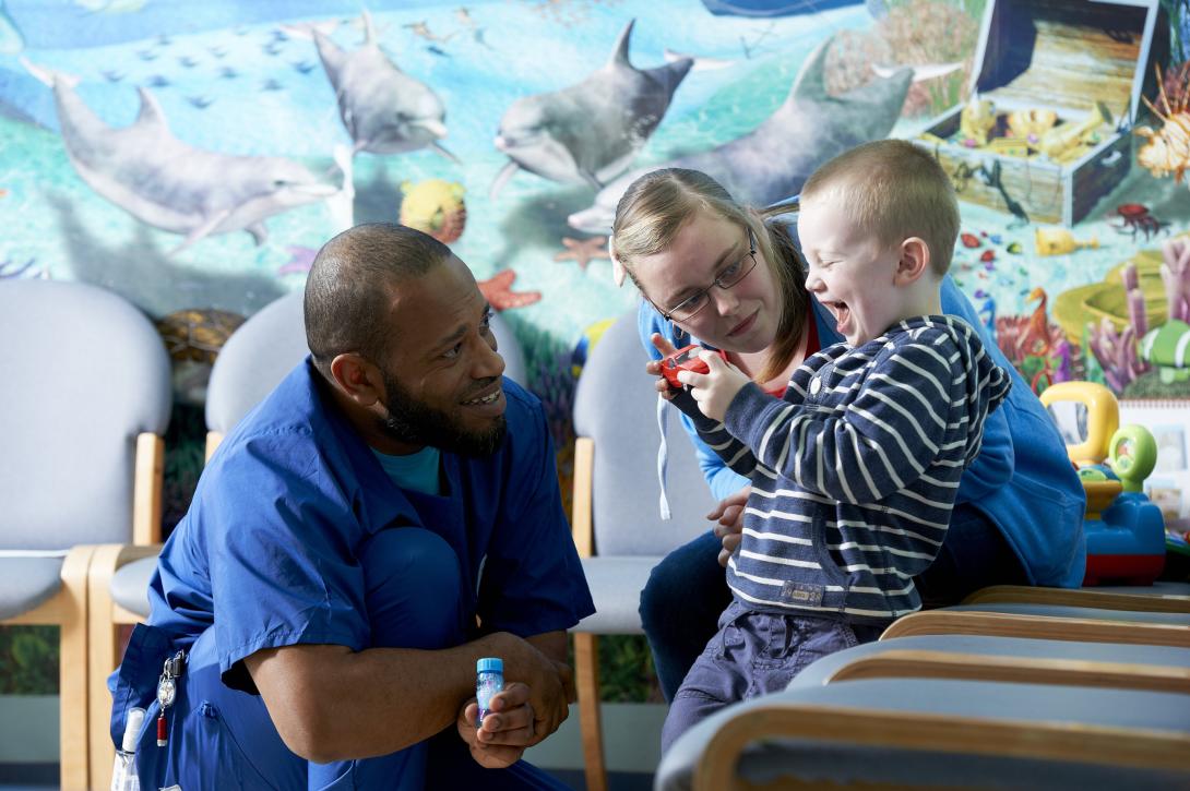 A nurse who is a Black man smiles at a young boy who is his patient; the boy is laughing. His mum sits beside him. 